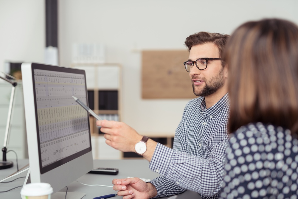 Proficient-young-male-employee-with-eyeglasses-and-checkered-shirt-explaining-a-business-analysis-displayed-on-the-monitor-of-a-desktop-pc-to-his-female-colleague-in-the-interior-of-a-modern-office - Developing Talent at the Bottom of the Corporate Ladder
