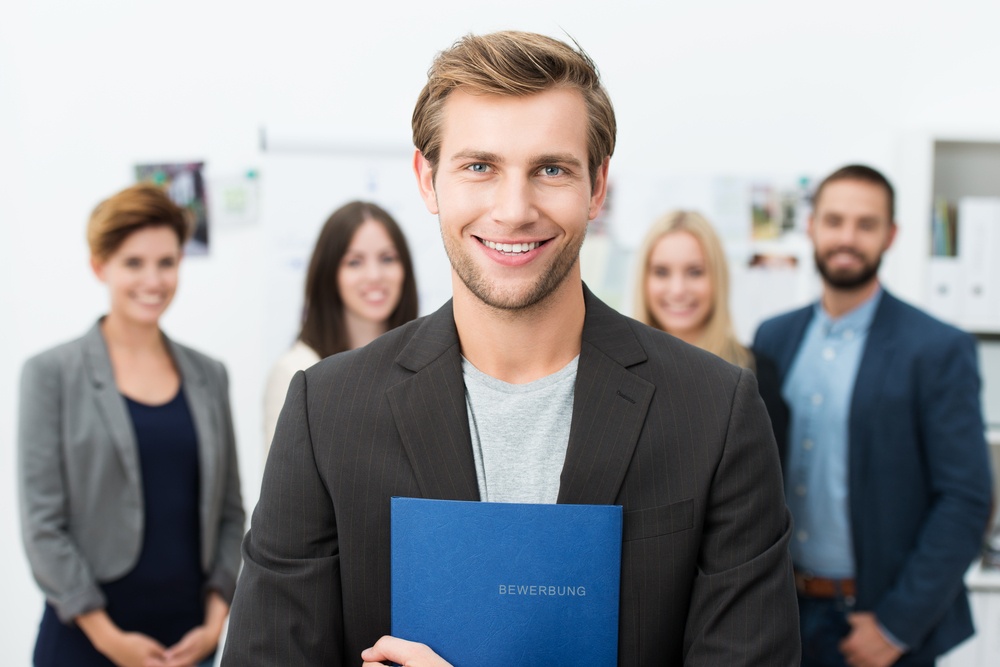 Successful-smiling-young-male-job-applicant-holding-a-blue-file-with-his-curriculum-vitae-posing-in-front-of-his-new-work-colleagues-or-business-team - Listening: The Key to Effective Business Relationships